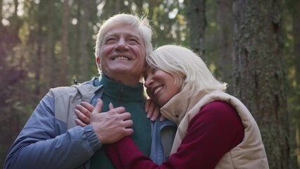 Senior elderly couple stay in sunny forest, hugging and enjoying leisure together. Tender love of old married couple embraces during stroll through the forest, smiling and admiring nature. Retirement