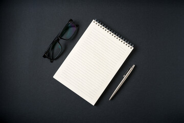 Top view of notebook, glasses and ballpoint pen on dark grey background