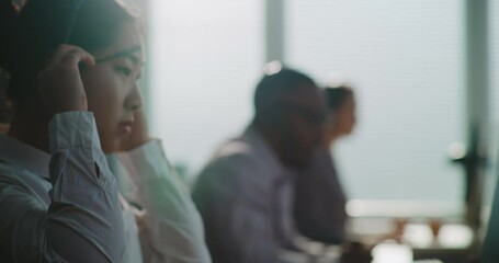 Modern call center office: Close up of Asian female technical support operator sitting down at computer, putting on headset. Multiethnic team of hotline specialists providing online customer service.