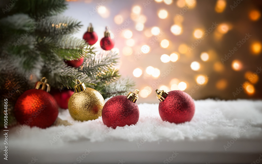 Poster red and gold christmas ornaments rest in snow with a pine branch and bokeh lights in the background.