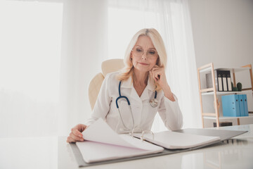 Senior female doctor reviewing medical records in a clinic office, highlighting dedication to...