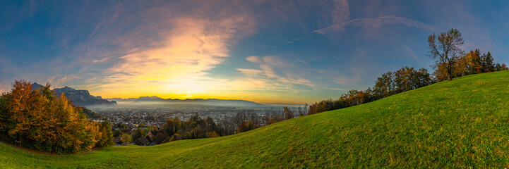 Panorama Sunset in the Rhine Valley, with a tree, blue sky over the city of Dornbirn, meadow and fields. autumn colored trees. beautiful afterglow, interesting colored veil clouds over Swiss mountains