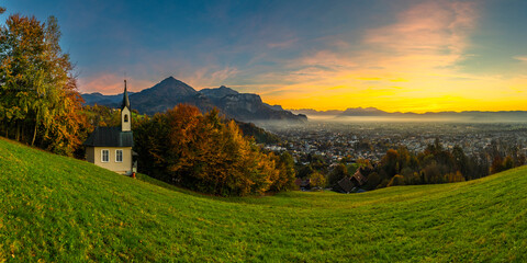 Obraz premium Panorama Sunset in the Rhine Valley, with a tree and a chapel,blue sky over the city of Dornbirn, meadow and fields. with Swiss mountains in the background. afterglow, interesting colored veil clouds