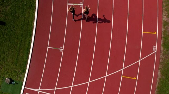 Overhead view of two female athletes running side by side on a red running track, shadows visible on the ground