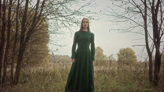 Model stands in a field surrounded by trees, wearing a long green dress. She is posing for the camera in a natural setting