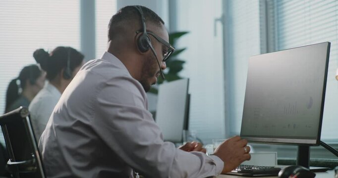 Modern call center office: Portrait of African American technical customer support specialist talking on headset, working on computer, solving problem, providing online help. Team of hotline operators