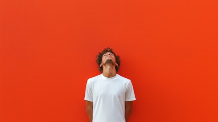 Man in white t-shirt standing against vibrant orange background looking up with a contemplative expression