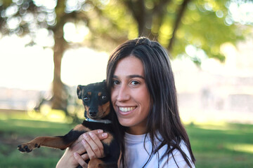 Smiling young woman embracing her pinscher dog in a park, capturing a moment of joy and...