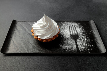 The sand basket is decorated with a high peak of protein cream. A tasty and sweet dessert stands on a black plate on a gray background
