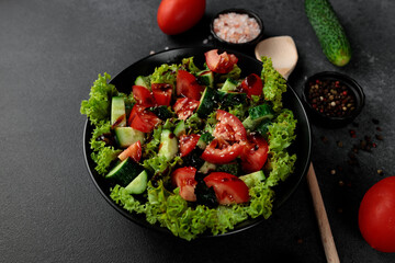 Vegetable salad of tomatoes, cucumbers, onions, lettuce dressed with aromatic dressing in a black plate on a gray background