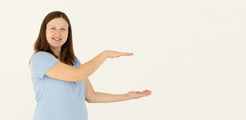 Young confident woman showing logo on empty space between arms. Smiling girl inviting or advertising promo deal, pointing hands right and looking at camera, white background.