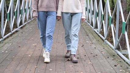 Two girlfriends are walking outside in autumn. View of the feet of two girls in autumn shoes walking along a wooden bridge in a city park.