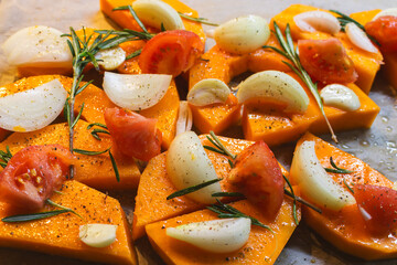 Sliced pumpkin with onion, garlic and rosemary leaves on baking tray. Autumn food still life. Raw pumpkin slices with herbs and spices, close up. Healthy eating concept. Vegan food. Dieting cooking.