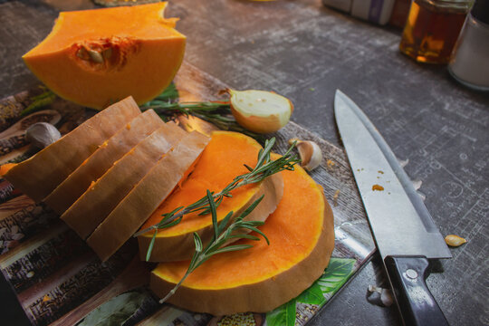 Sliced pumpkin with onion, garlic and rosemary leaves on cutting board. Autumn food still life. Raw pumpkin slices with herbs and spices and knife, close up. Healthy eating concept. Vegan food. 
