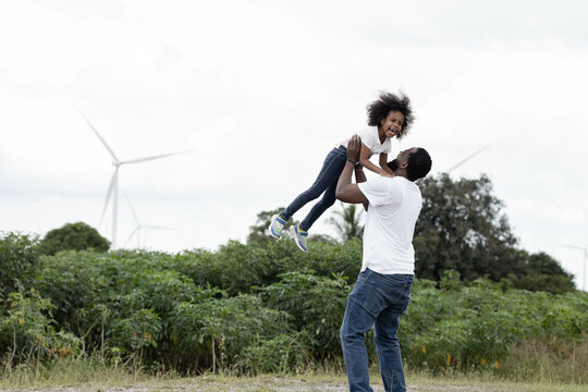 A black father is teasing his daughter by using both arms to lift her above his head. Father and daughter are smiling and their happy in the nature in windmill.