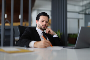 A man in a suit is writing on a piece of paper with an orange pen. He is deep in thought, possibly...