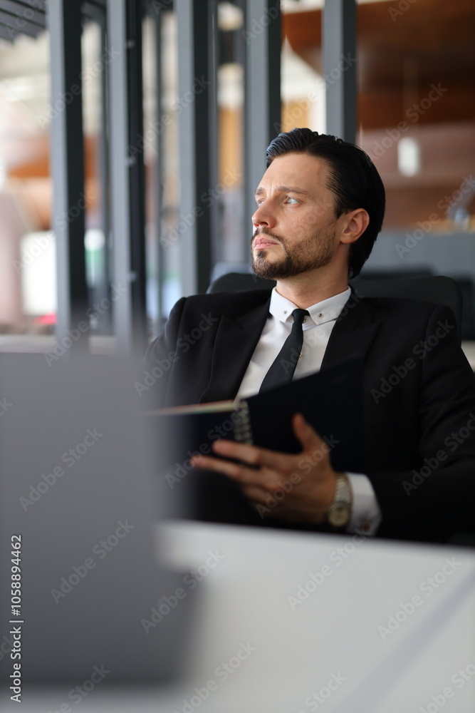 Poster a man in a suit is sitting at a desk with a laptop and a notebook. he is looking at the notebook and