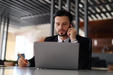 A man in a suit is talking on his cell phone while looking at a laptop. He is wearing a tie and he is in a professional setting