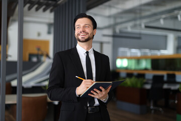 A man in a suit is smiling and holding a pen and a notebook. He is happy and focused on writing something