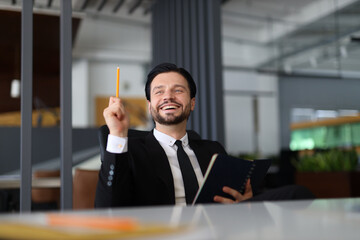 A man in a suit is holding a pencil and smiling. He is sitting at a desk with a book in front of...