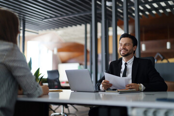 A man in a suit is sitting at a desk with a woman. He is smiling and holding a piece of paper. The woman is sitting across from him, looking at him