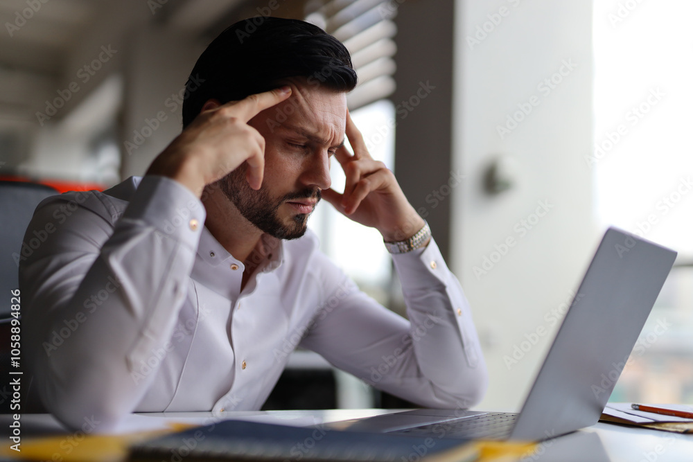 Poster a man is sitting at a desk with a laptop open in front of him. he is looking at the screen with a fu