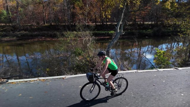A mature woman in a green tank top and black shorts cycles on a paved path near a river. The trees along the river are changing colors, indicating autumn.