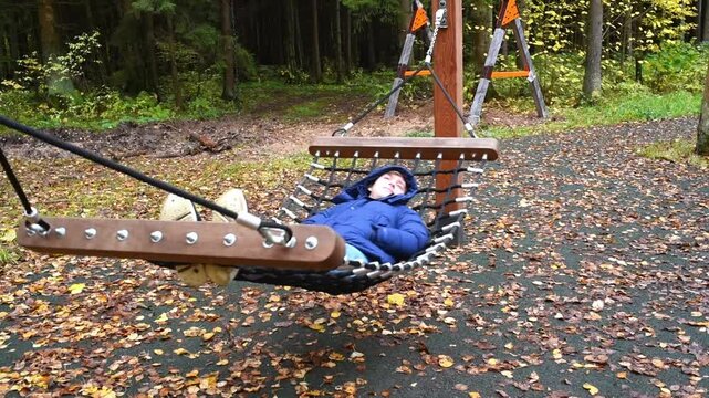 A boy in a blue jacket and jeans lies and rests in a hammock on a playground in the autumn Borovsky Park in Moscow.