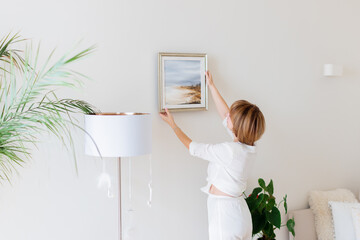Woman hanging picture on wall in room. Interior design.