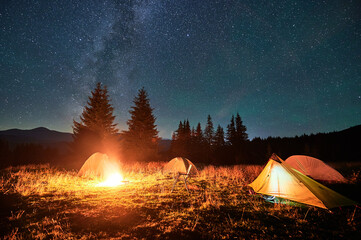 Several tents around glowing campfire on grassy clearing under starry sky with Milky Way. Warm...
