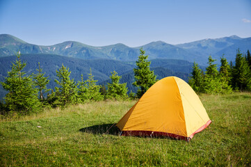 Bright yellow tent on grassy hillside, overlooking breathtaking panorama of blue-tinged mountains. Young pine trees under clear blue sky, creating serene and picturesque camping spot.
