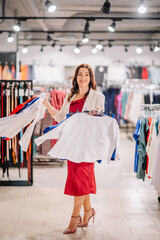 Woman in red dress and beige blazer holding colorful shirts while shopping in a stylish clothing store, looking happy and confident..