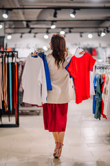  Rear view of a woman holding red, blue, and white shirts in a clothing store, deciding on her purchase. Bright, spacious store with copy space.