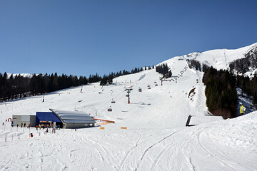 Panorama view of winter ski resort with buildings and ski track with snowboarders and skiers. Blue sky above the mountain. Winter sports and recreation