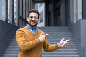 A young man stands near an office building and points with his fingers to an empty space nearby, looks and smiles at the camera