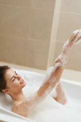 Relaxed young woman enjoying a bubble bath, smiling peacefully while applying foam to her arms....