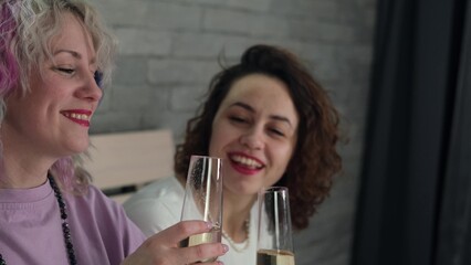 Two women with curly hair and bright makeup drink champagne in the bedroom at home and have fun