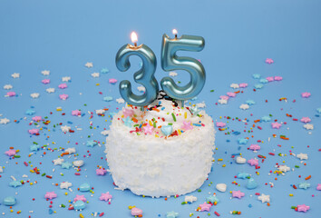 Coconut birthday cake on blue background with burning candles number 35 and many colorful sprinkles.