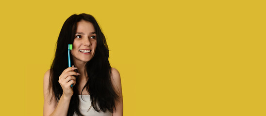 Fototapeta premium A smiling brunette woman with healthy teeth holds a toothbrush in her hands. A young beautiful girl on a bright background. Oral hygiene. A place for your text.