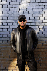 Obraz premium African american man in sunglasses and leather jacket posing near brick wall 
