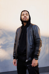 Young african american man in hoodie and leather jacket standing near wall with sunlight