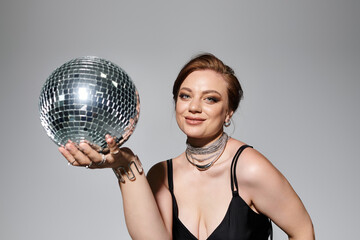 Fototapeta premium A beautiful young woman, dressed elegantly, playfully holds a sparkling disco ball, exuding charm.