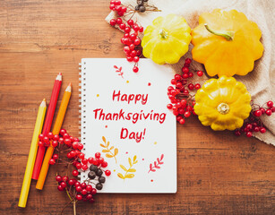 Happy Thanksgiving Day! White notebook with colored pencils, yellow squash with red viburnum berries on old wooden surface, top view.