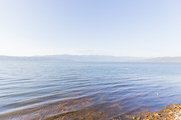 Tranquil Scene Of Doirani Lake With Its Calm Waters Stretching To The Horizon, Framed By Distant Mountain Ranges And A Clear, Blue Sky