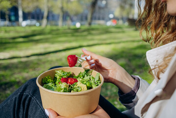 Young lady delights in a nourishing lunch at the park, relishing a crisp salad with sustainably sourced strawberries in a to-go bowl