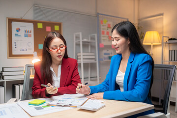 Two women in suits are sitting at a desk with a white board behind them