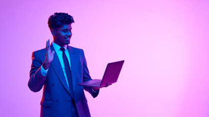 Portrait of young smiling Indian man in formal wear holding laptop and waving into screen, greeting with colleagues against purple studio background in neon light. Concept of emotions, lifestyle