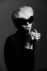 Stylish woman with short white hair and oversized sunglasses, striking a moody pose with hands clasped, set against a dark monochrome background.