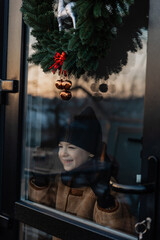 a portrait of a cute girl looking through the glass of a Christmas decorated door in anticipation of the holiday