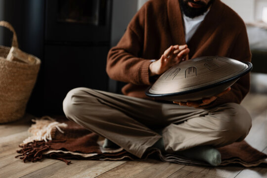 young bearded man with a melodious musical instrument that emits cosmic sounds: glucophone or handpan, stylish photo of playing tongdrum in the interior of a white wooden house.​